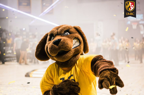 True Grit mascot looking at camera and pointing with gold and black confetti in background.