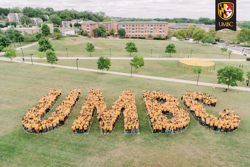 Overhead shot of large group of students wearing gold shirts and standing in groups to spell out U M B C.