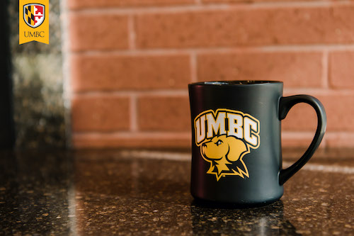Black UMBC mug with gold Retriever logo sitting on a table.