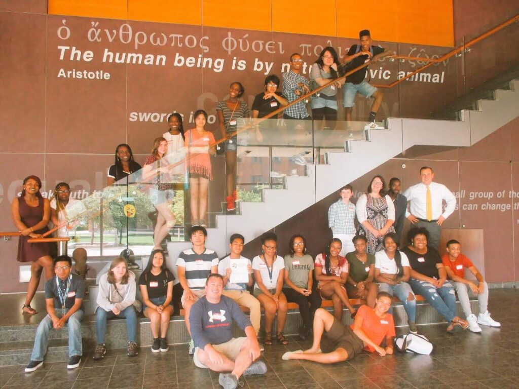Over two dozen young people stand on a staircase and on a step inside a building, posing for a portrait. Text on the wall behind them reads, 