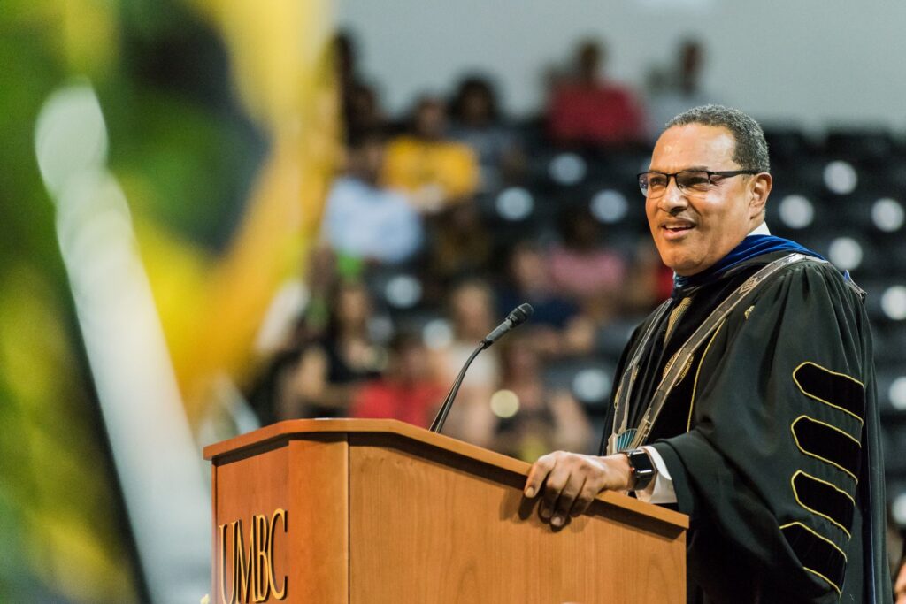 Man stands at podium in full academic regalia. Podium sign reads