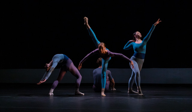 Four dancers perform a contemporary routine on a dark stage, executing synchronized leaps, arches, and extensions.