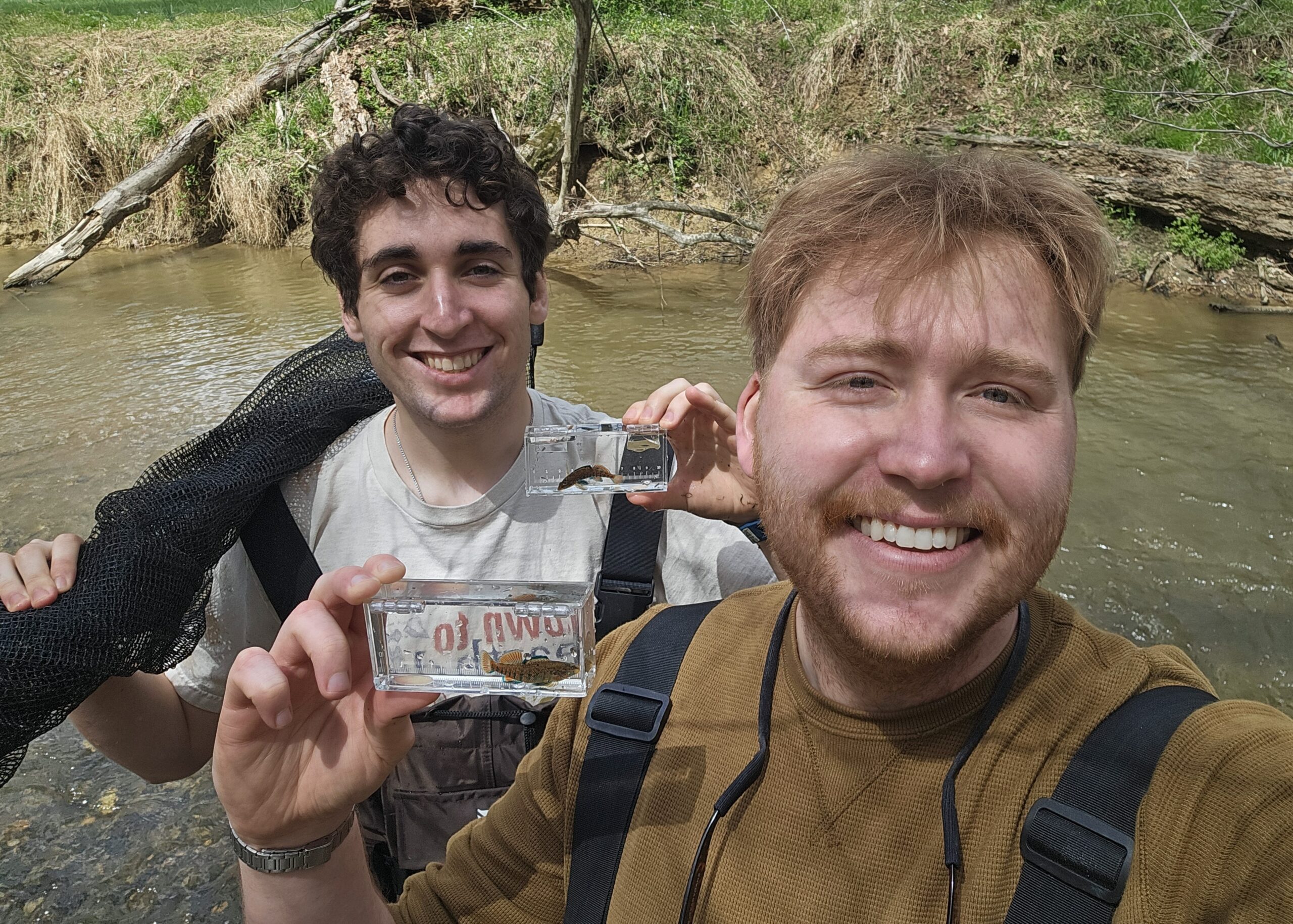 two people holding clear plastic containers each holding a fish, backed by a stream scene