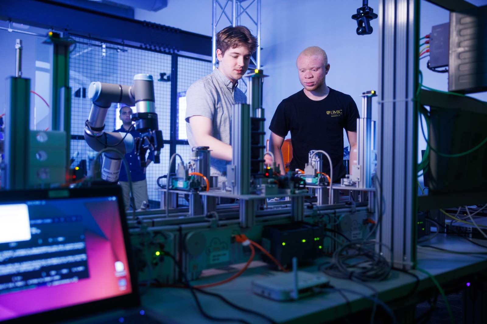 Two students work with a robotic arm in an engineering lab. A computer monitor is in the foreground.