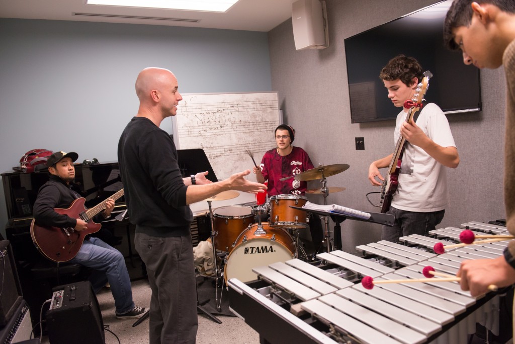 A music instructor directs students playing electric guitars, drums, and a vibraphone in a practice room. A television is mounted on the wall.
